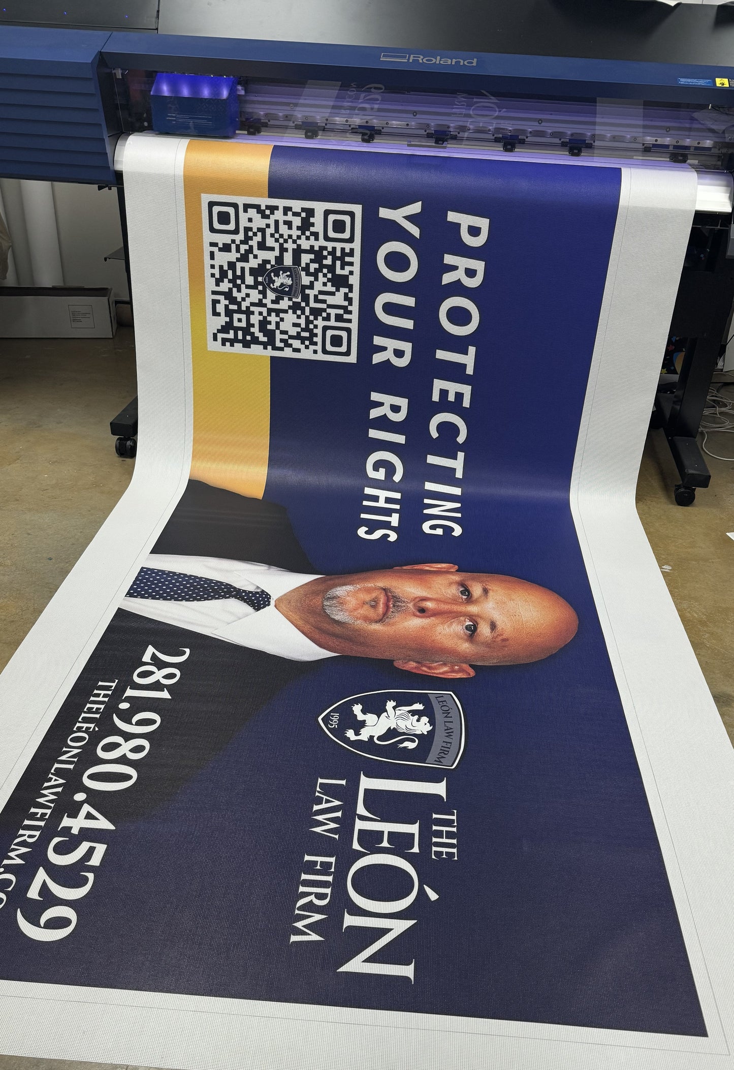 A large promotional banner featuring a bald man in a suit, with text "protecting your rights" and "the leon law firm" coming out of a Show Off Your Threads Custom Mesh Indoor/Outdoor Banner at a printshop, including.