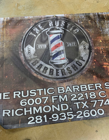 A weathered Custom Mesh Indoor/Outdoor Banner for "the rustic barbershop," featuring a logo with a barber pole, established in 2022, on a metallic background with custom embroidery and contact details for a location from Show Off Your Threads.