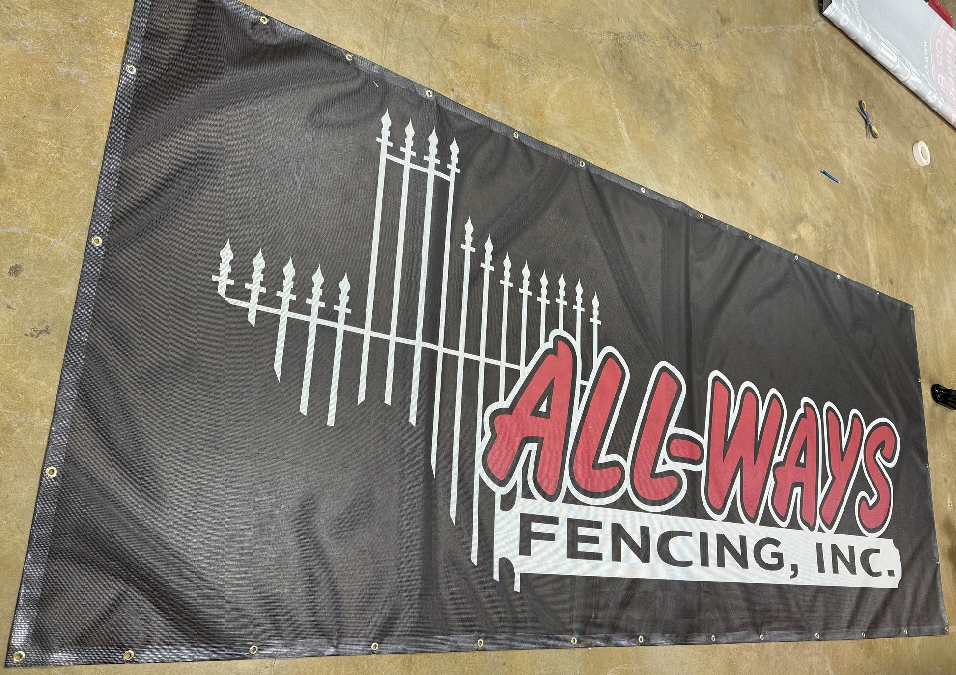 A large Show Off Your Threads custom mesh indoor/outdoor banner for "allways fencing, inc." featuring a stylized white fence design on a dark background, displayed on a workshop floor with visible tools.