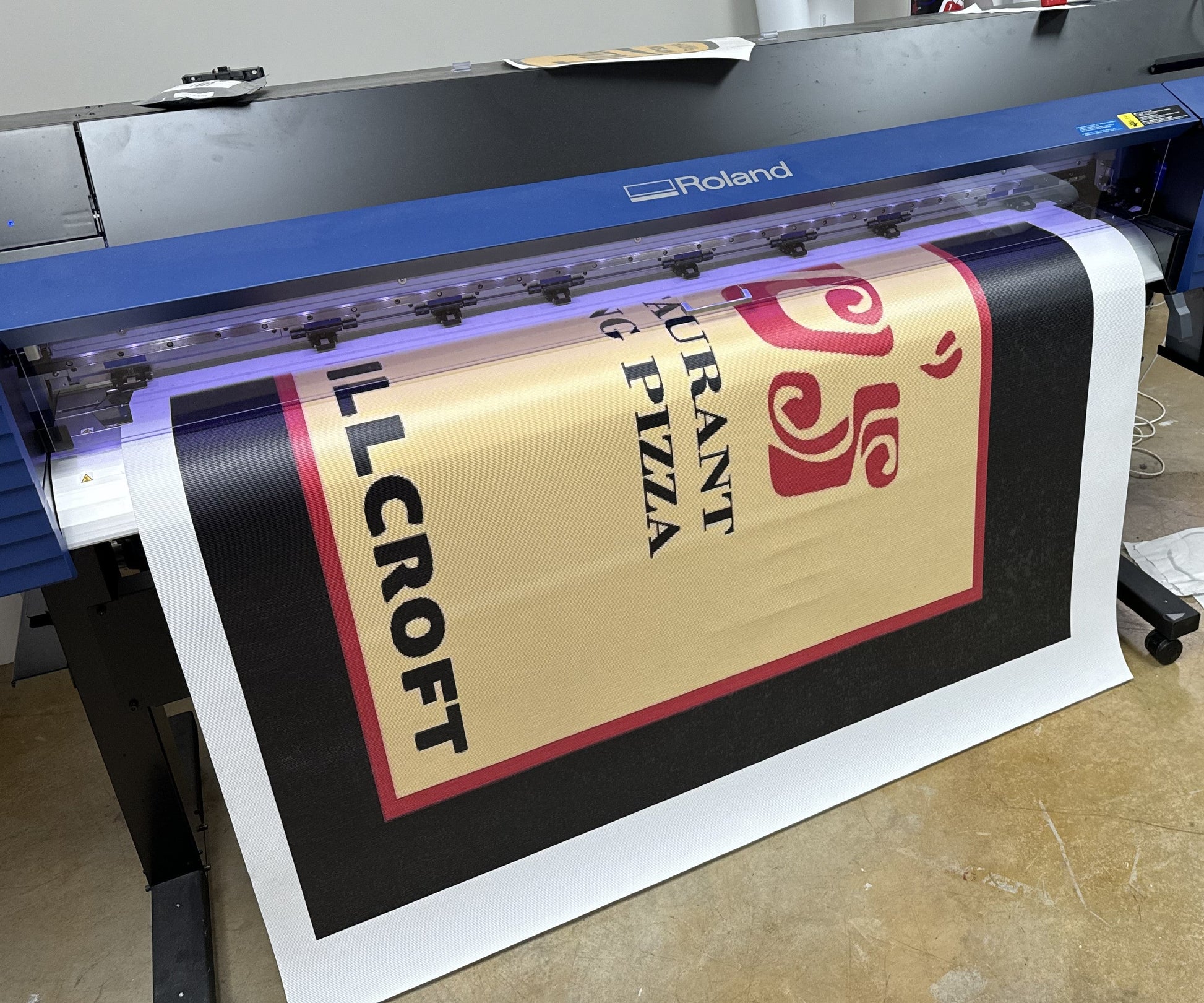 Custom Mesh Indoor/Outdoor Banner by Show Off Your Threads custom printed for "Grant's Pizza" on a Roland digital printer in a printshop workspace. The banner is yellow with red and black text detailing the business name.